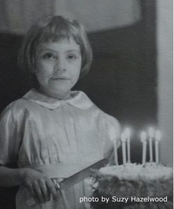 a black and white image of a young white girl wearing a dress, standing with a knife behind a cake topped by burning candles